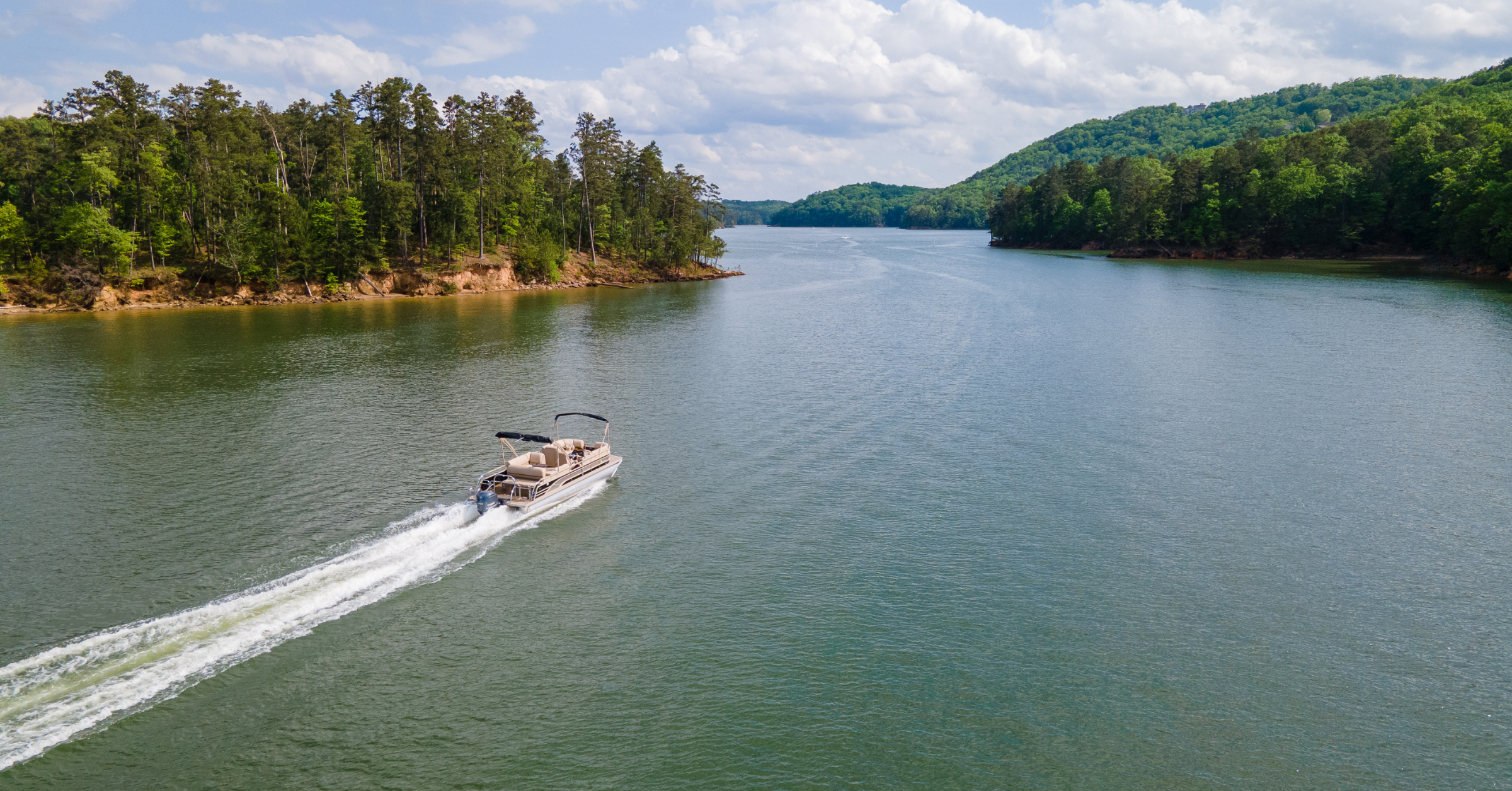 Pontoon boat cruising across a scenic lake surrounded by green hills, highlighting the perfect setting for a lake day with must-have pontoon boat accessories.